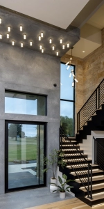 Large glass pivoting front door with quartz wall and cieling panels with pendant lighting design next to black staircase.